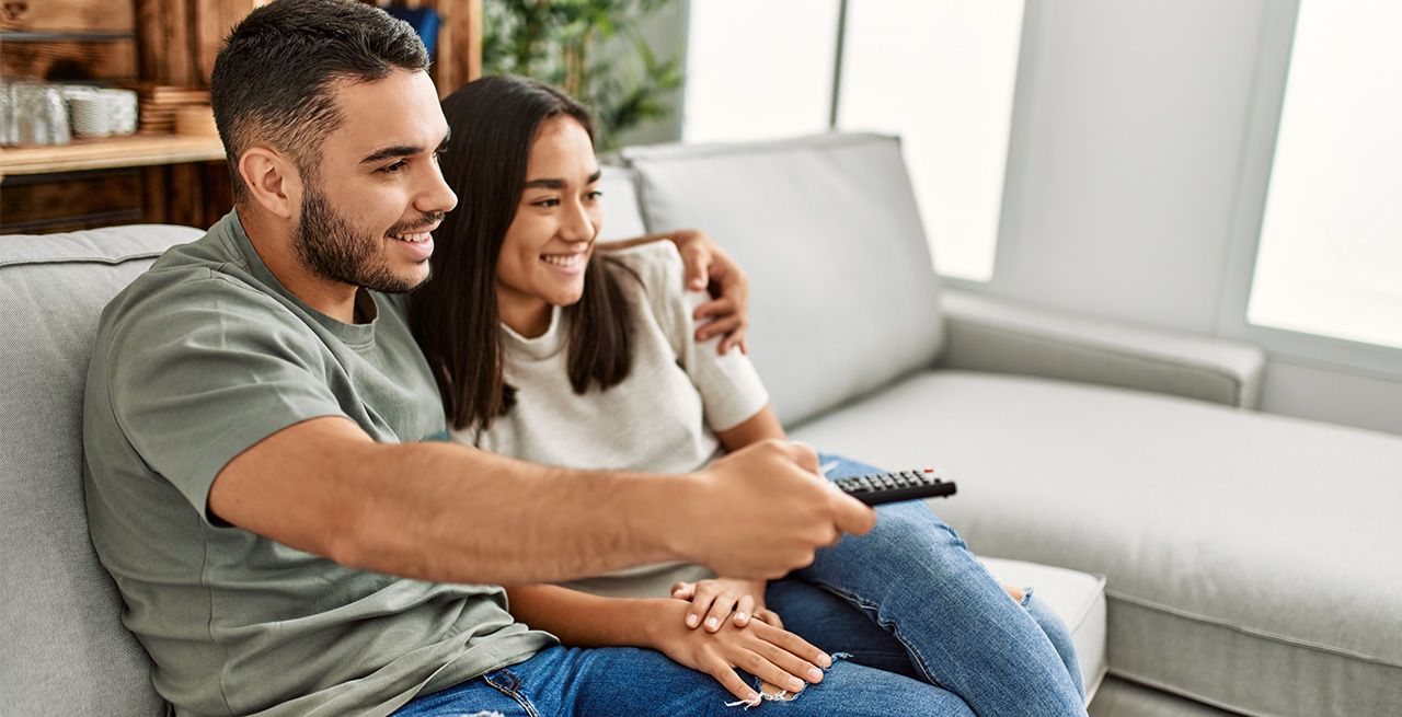 Man and woman possibly married watching TV from their living room couch, man has remote in his hand 