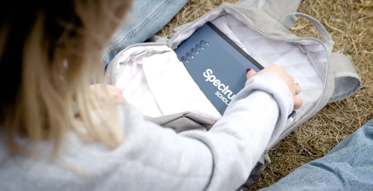 College student sitting on grass looking through her backpack with Spectrum-branded notebook inside