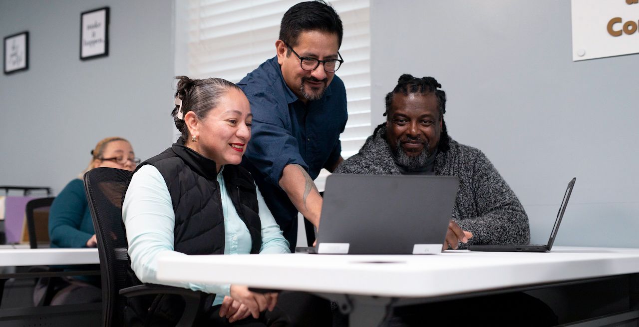 Computer lab instructor helping two adult students on their laptops 