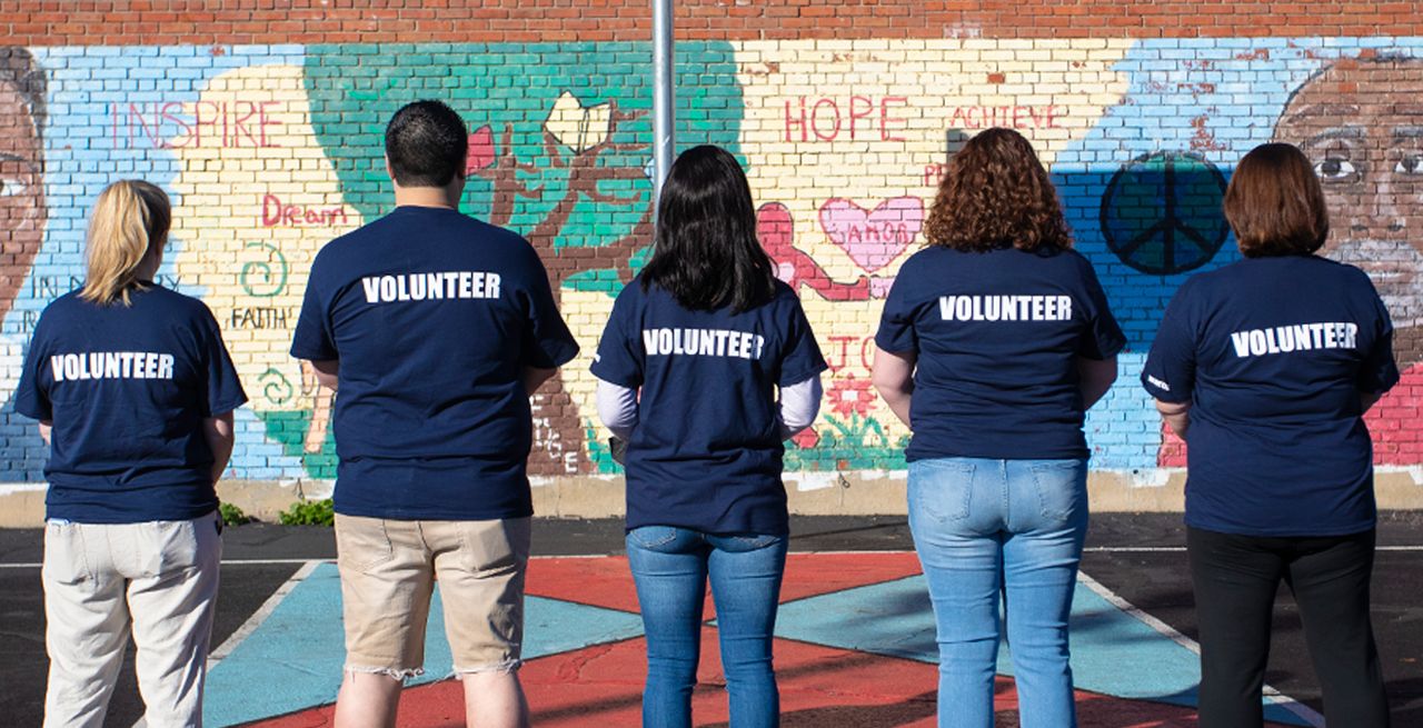 Spectrum volunteers on the grounds of a community center, standing in a row, backs turned, with "Volunteer" t-shirts on
