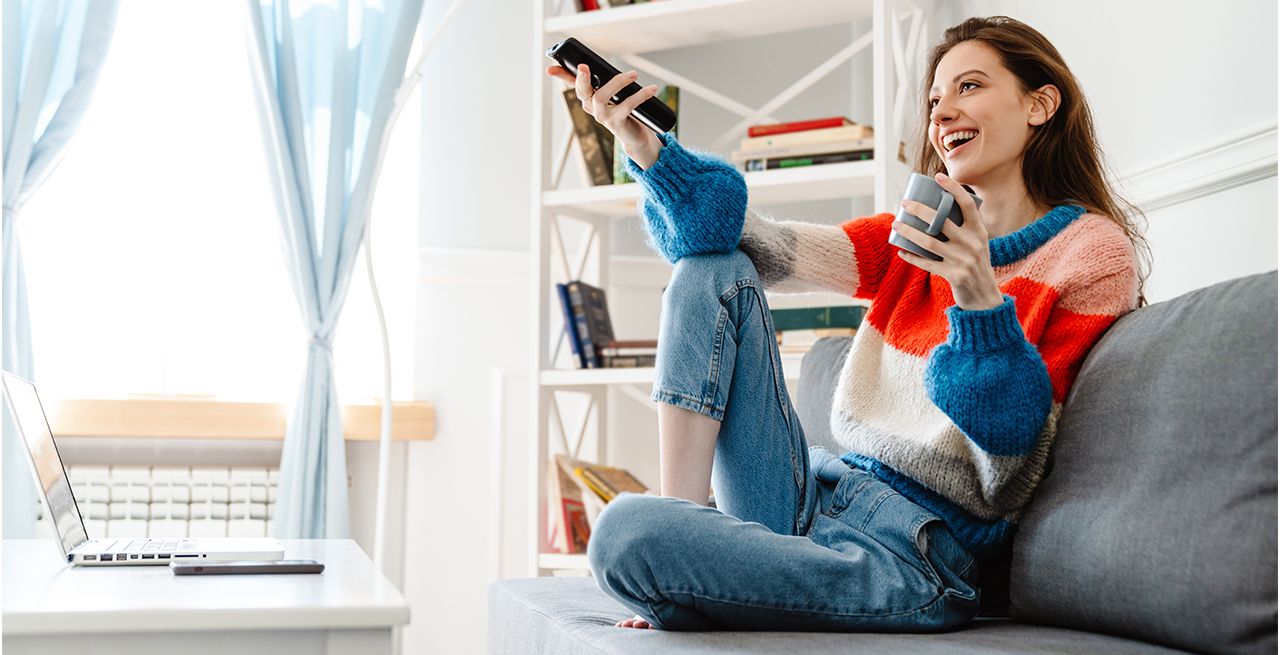 Young woman holding a remote watching TV from her couch, smiling happily, with coffee