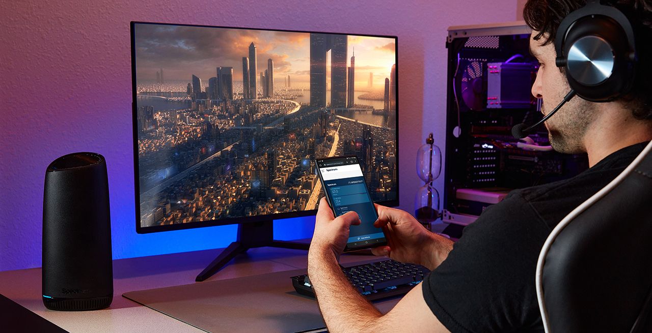A Spectrum customer using his mobile phone while sitting at his desk with WiFi 7 router and desktop computer on the desk
