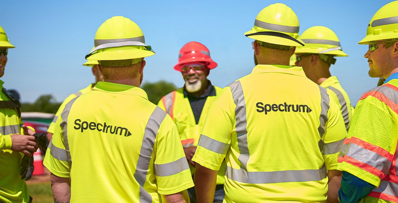 A team of about 6 or 7 Spectrum field technicians standing in a circle, during a planning session, yellow hard hats and Spectrum-branded vests on