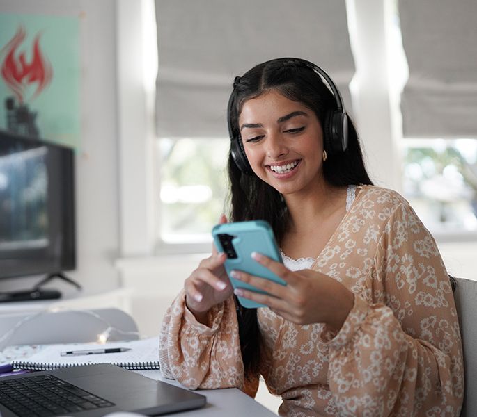 A young woman using her smartphone in her room, at her desk, headphones on, smiling