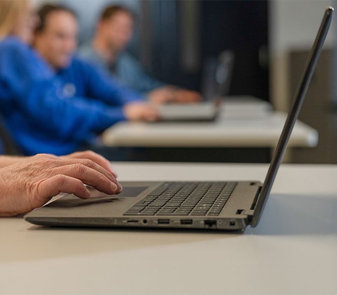 Someone using a laptop in a computer lab, hands showing only, with adult students blurred in the background