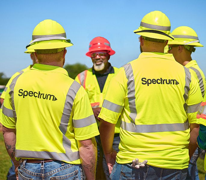 A team of Spectrum field technicians standing in a circle for a team meeting, in yellow hard hats and vests