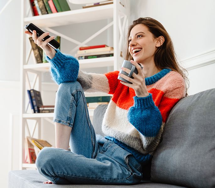 A young woman using her TV remote on her couch at home, with coffee, smiling